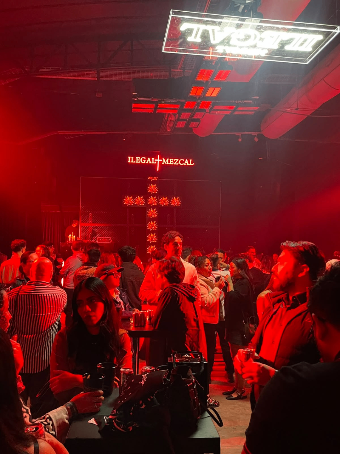 Fiesta Ilegal audience beneath the central red lighting installation, with the adapted stage visible in the final venue.