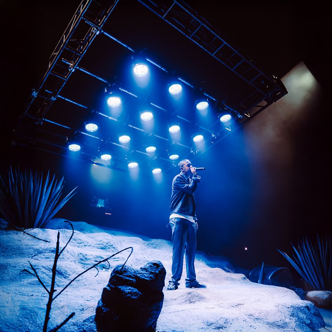 Live event photograph: Natanael Cano performing on the built dune under blue light, with relief and desert vegetation visible